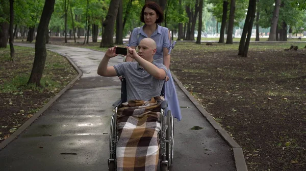 Eine Frau führt einen jungen Mann mit Onkologie im Rollstuhl durch den Park. Ein Mann sitzt mit einem Handy in der Hand und macht Fotos von der umgebenden Natur. Haarausfall durch Chemotherapie. — Stockfoto