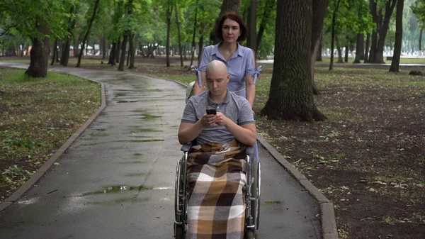 女性は車椅子の公園で腫瘍学の若い男をリードしています。男は携帯電話を手にして座っている。化学療法による脱毛. — ストック写真