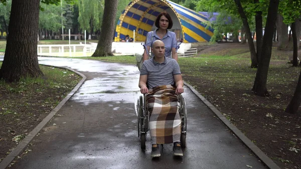 Μια γυναίκα οδηγεί έναν νεαρό με ογκολογικό σε αναπηρικό καροτσάκι στο πάρκο. Ο άνθρωπος είναι φαλακρός λόγω χημειοθεραπείας.. — Φωτογραφία Αρχείου