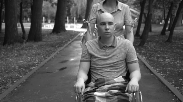 Una mujer está conduciendo a un hombre joven con oncología en silla de ruedas a través del parque. El hombre es calvo debido a la quimioterapia. Color monocromo. —  Fotos de Stock