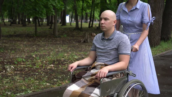 Una mujer está conduciendo a un hombre joven con oncología en silla de ruedas a través del parque. El hombre es calvo debido a la quimioterapia.. — Foto de Stock
