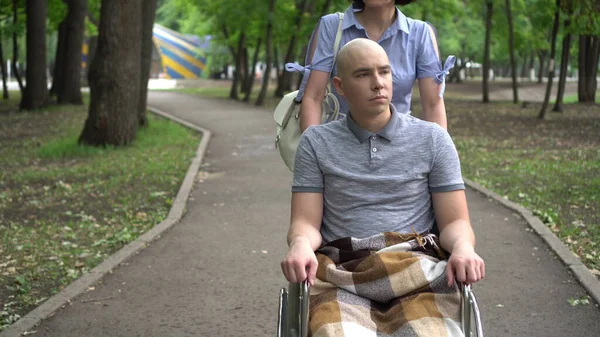 Uma mulher está dirigindo um jovem com oncologia em uma cadeira de rodas através do parque. O homem é careca devido à quimioterapia.. — Fotografia de Stock