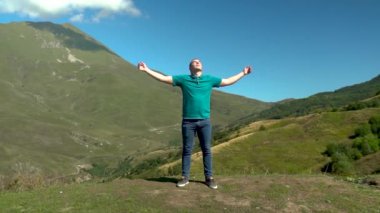 Genç bir adam kollarını dağların arka planına kaldırmış bir elbiseyle ayakta duruyor. Yavaş çekim.