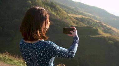 Elbiseli genç bir kadın dağlarda dikiliyor ve telefonda fotoğraf çekiyor. Yavaş çekim.
