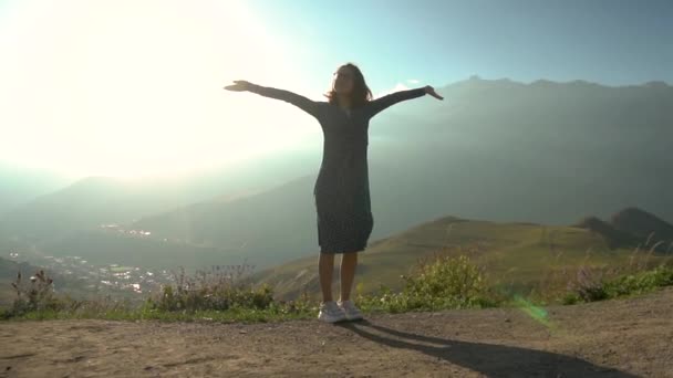 Een jonge vrouw staat in een jurk en steekt haar handen omhoog kijkend naar de bergen. Langzame beweging. — Stockvideo
