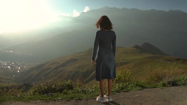 En ung kvinna står i klänning och sträcker upp händerna och tittar på bergen. Långsamma rörelser. — Stockvideo