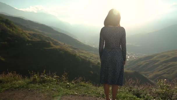 Une jeune femme se tient dans une robe et lève les mains vers le haut en regardant les montagnes. Mouvement lent. — Video