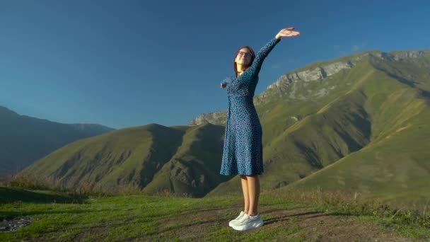 Une jeune femme se tient en robe, les bras levés sur fond de montagnes. Mouvement lent. — Video