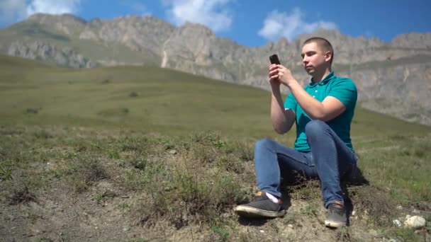Ένας νεαρός άνδρας κάθεται με ένα τηλέφωνο στα χέρια του στα βουνά. Ο άντρας είναι στην αλληλογραφία του smartphone στα βουνά του Καυκάσου.. — Αρχείο Βίντεο