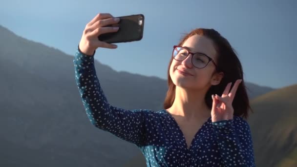 Elbiseli genç bir kadın dağlarda dikiliyor ve akıllı telefondan selfie çekiyor.. — Stok video