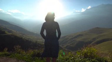 Genç bir kadın elbise giymiş ve ellerini kaldırıp dağlara bakıyor. Güneş dağların kenarından ışıl ışıl parlıyor.