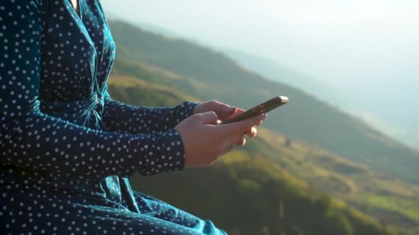 Una joven con un vestido se sienta con un teléfono en sus manos sobre un fondo de montañas de cerca. La chica viaja por las montañas del Cáucaso. — Vídeos de Stock