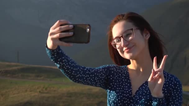 En ung kvinna i klänning står i bergen och tar en selfie på en smartphone. Flickan reser i de vita bergen. — Stockvideo