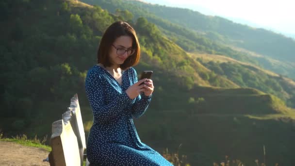 Una joven con un vestido se sienta en un banco con un teléfono en sus manos sobre un fondo de montañas. La chica viaja por las montañas del Cáucaso. — Vídeos de Stock