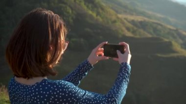 Elbiseli genç bir kadın dağlarda dikiliyor ve telefonda fotoğraf çekiyor. Kız Kafkas dağlarında seyahat ediyor..