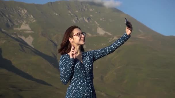 Μια νεαρή γυναίκα με φόρεμα στέκεται στα βουνά και βγάζει μια σέλφι σε ένα smartphone.. — Αρχείο Βίντεο