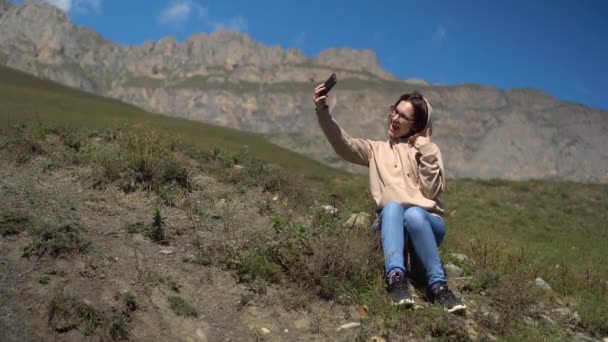 Egy fiatal nő pulóverben ül a hegyekben és szelfit készít egy okostelefonon. A lány a kaukázusi hegyekben utazik.. — Stock videók