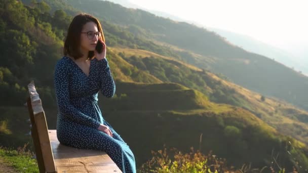 Uma jovem mulher em um vestido senta-se em um banco e fala ao telefone contra um fundo de montanhas. A menina viaja nas montanhas caucasianas. — Vídeo de Stock