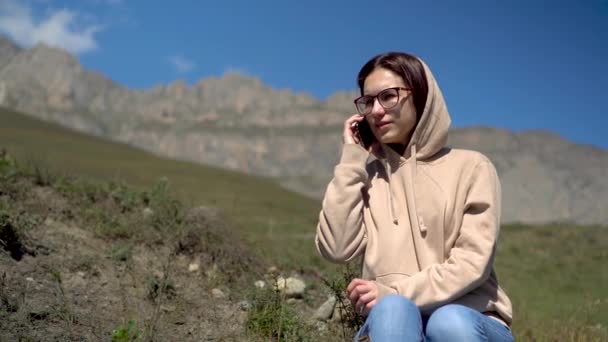 Een jonge vrouw in een sweater zit in de bergen en spreekt aan de telefoon. Het meisje reist in de Kaukasische bergen. — Stockvideo