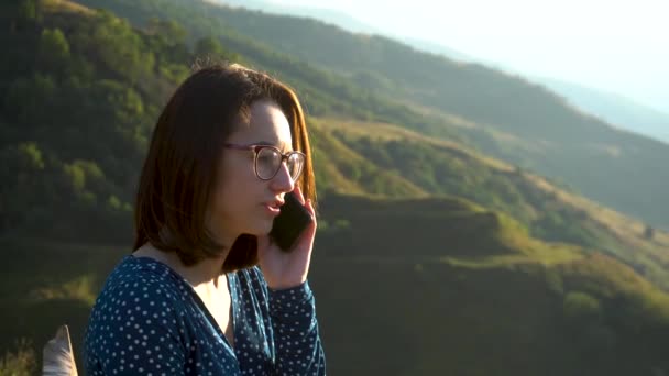 Une jeune femme en robe s'assoit et parle au téléphone sur fond de montagnes. La fille voyage dans les montagnes du Caucase. — Video