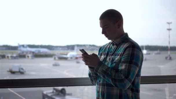 Молодой человек с телефоном в руках на фоне окна в аэропорту. Самолеты на заднем плане. — стоковое видео