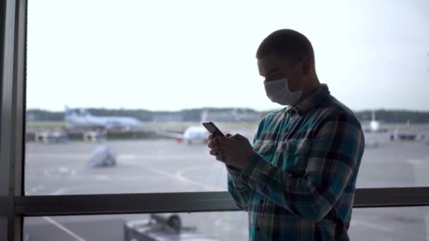 의료용 마스크를 쓴 채 손에 전화기를 들고 공항 창문을 등지고 있는 한 젊은이. — 비디오