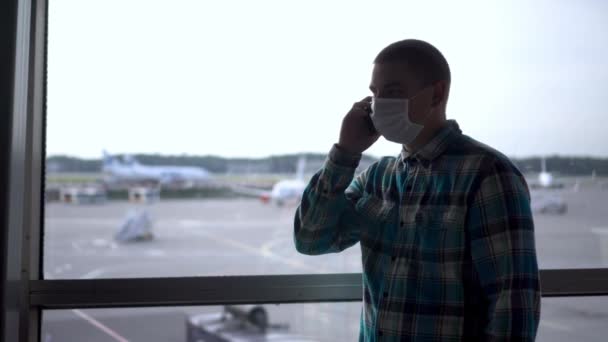 Un giovane con una maschera medica parla al telefono sullo sfondo di una finestra all'aeroporto. Aerei sullo sfondo. — Video Stock