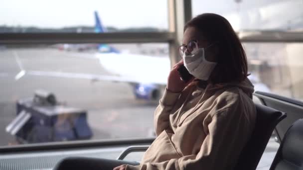 若い女性が空港の窓の後ろに座って電話で話します。背景に飛行機. — ストック動画