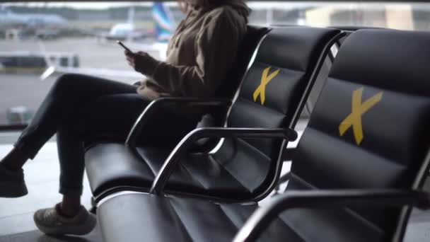 Uma jovem mulher com uma máscara médica senta-se com um telefone nas mãos contra o fundo de uma janela no aeroporto. As cadeiras são marcadas com cruzes para manter a distância social. — Vídeo de Stock