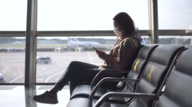 Tıbbi maskeli genç bir kadın, elinde telefonuyla havaalanındaki bir pencerenin arkasında oturuyor. Sandalyeler sosyal mesafeyi korumak için haçlarla işaretlenmiş..