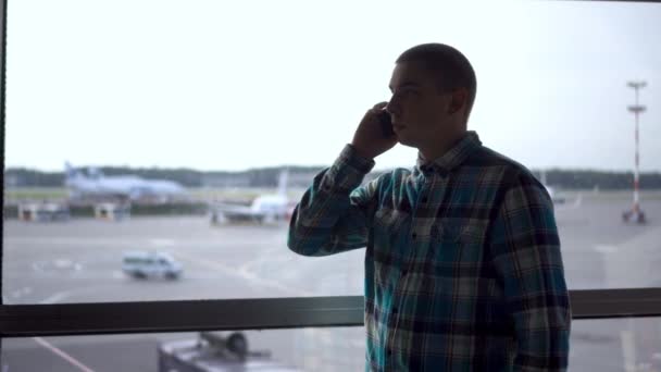 한 젊은이가 공항 창밖을 배경으로 전화 통화를 하고 있다. 뒤에 있는 비행기들. — 비디오
