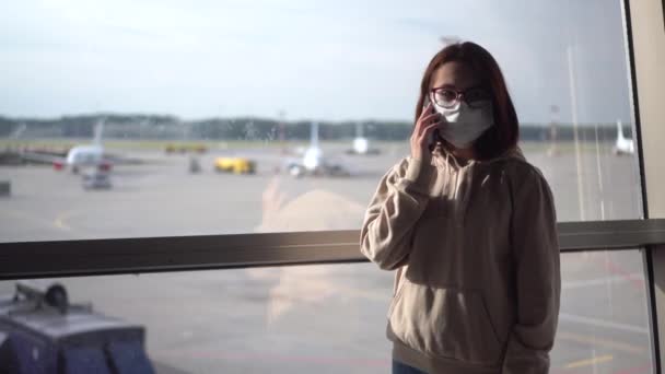 Młoda kobieta w masce medycznej rozmawia przez telefon na tle okna na lotnisku. Samoloty w tle. — Wideo stockowe
