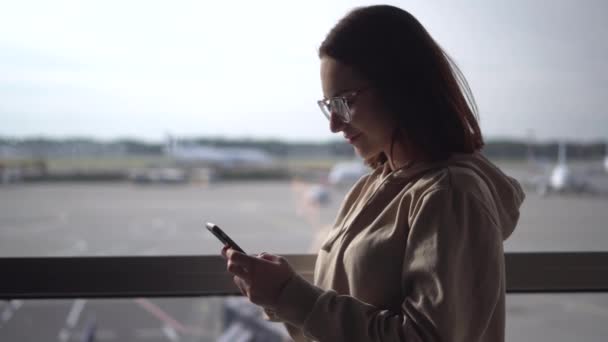 Jovem com um telefone em suas mãos no fundo de uma janela no aeroporto closeup. Aviões de fundo. — Vídeo de Stock