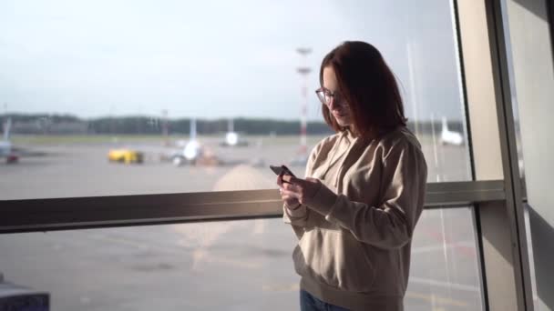 Jovem com um telefone nas mãos no fundo de uma janela no aeroporto. Aviões de fundo. — Vídeo de Stock