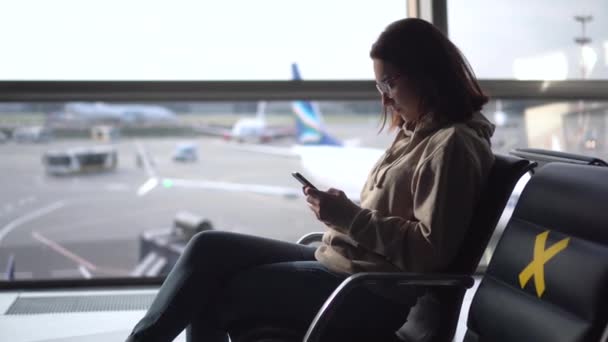 Молодая женщина сидит с телефоном в руках на фоне окна в аэропорту. Стулья отмечены крестами, чтобы сохранить социальную дистанцию. — стоковое видео