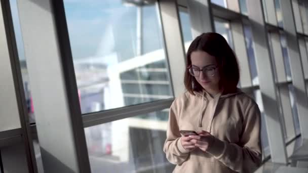 Молодая женщина в толстовке и очках идет по аэропорту с телефоном в руках на фоне окна. — стоковое видео