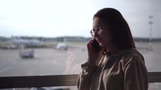 Une jeune femme en sweat-shirt et lunettes parle au téléphone sur le fond d'une fenêtre près de l'aéroport. Avions en arrière-plan. — Video