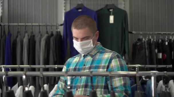 A young man in a medical mask is choosing clothes in a store. Clothing store. — Stock Video