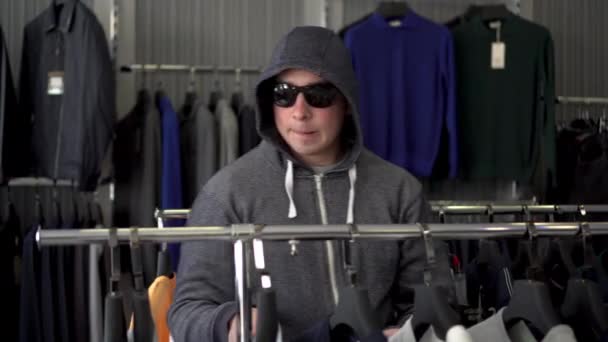 Un hombre con capucha y gafas roba una camiseta de una tienda. Tienda de ropa. — Vídeo de stock