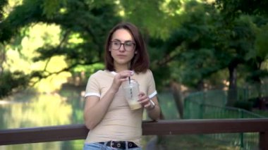 Gözlüklü genç bir kadın parkta bir köprüde duruyor ve soğuk kahve içiyor. Arka planda bir göl var..