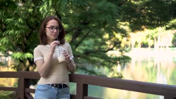 Młoda kobieta w okularach stoi na moście w parku i pije zimną kawę z bliska. W tle jest jezioro.. — Wideo stockowe