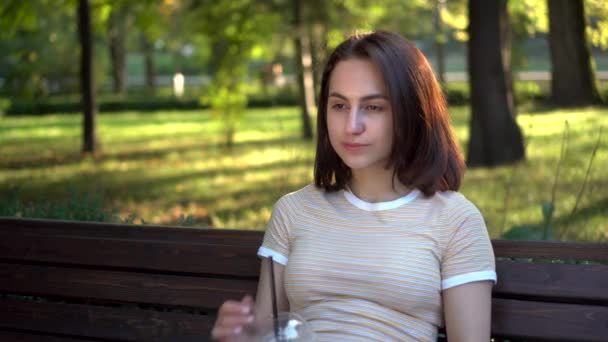 Een jonge vrouw met een bril zit op een bankje in het park en drinkt koude koffie close-up. Op de achtergrond is er een meer. — Stockvideo