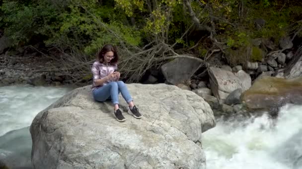 Egy fiatal lány ül egy kőre egy telefonnal a kezében a hegyekben a folyó mellett.. — Stock videók