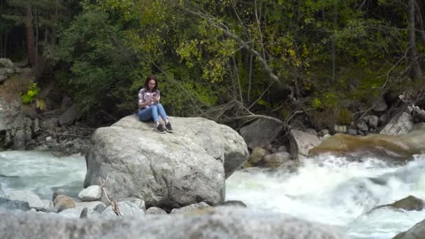 Egy fiatal lány ül egy kőre egy telefonnal a kezében a hegyekben a folyó mellett. Utazás a hegyekbe. — Stock videók