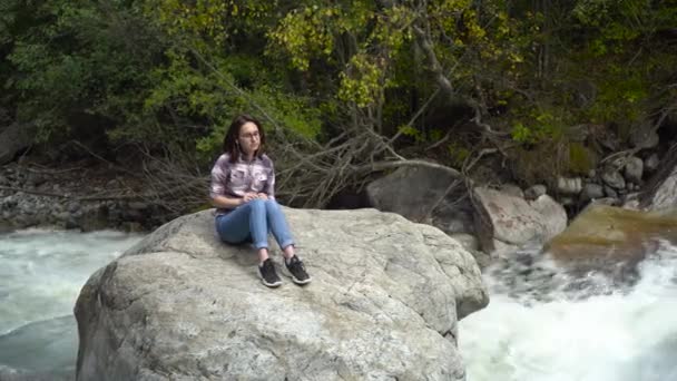 Egy szemüveges fiatal nő ül egy kőre fejhallgatóval a hegyekben a folyó közelében. Utazás a hegyekbe. — Stock videók