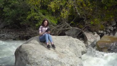 Gözlüklü genç bir kadın nehrin yanındaki dağlarda elinde tabletle bir taşın üzerinde oturuyor. Dağlara git..