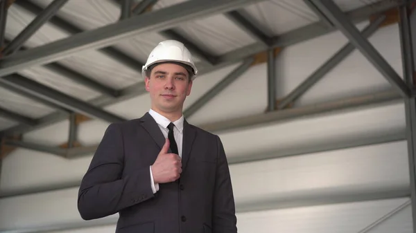 A young man in a protective helmet, shows a thumb at a construction site. The boss in a suit looks at the camera. — Stock Photo, Image