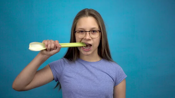 Dişlerinde diş teli olan genç bir kız mavi arka planda kereviz yiyor. Renkli telleri olan bir kız kereviz sapını ısırıp koparır..