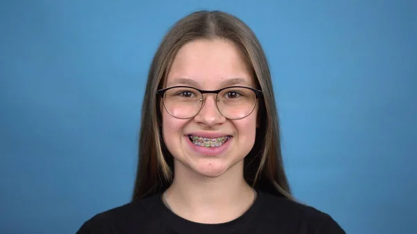 Nastolatka z aparatem na zębach uśmiechnięta na niebieskim tle. Dziewczyna z kolorowymi aparatami na zęby. — Zdjęcie stockowe