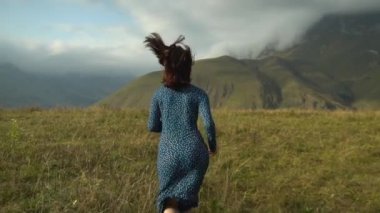 Mavi elbiseli genç bir kadın tepeden aşağı doğru koşuyor. Kafkasya bulutlu dağlarının arka planına doğru. Yavaş çekim.